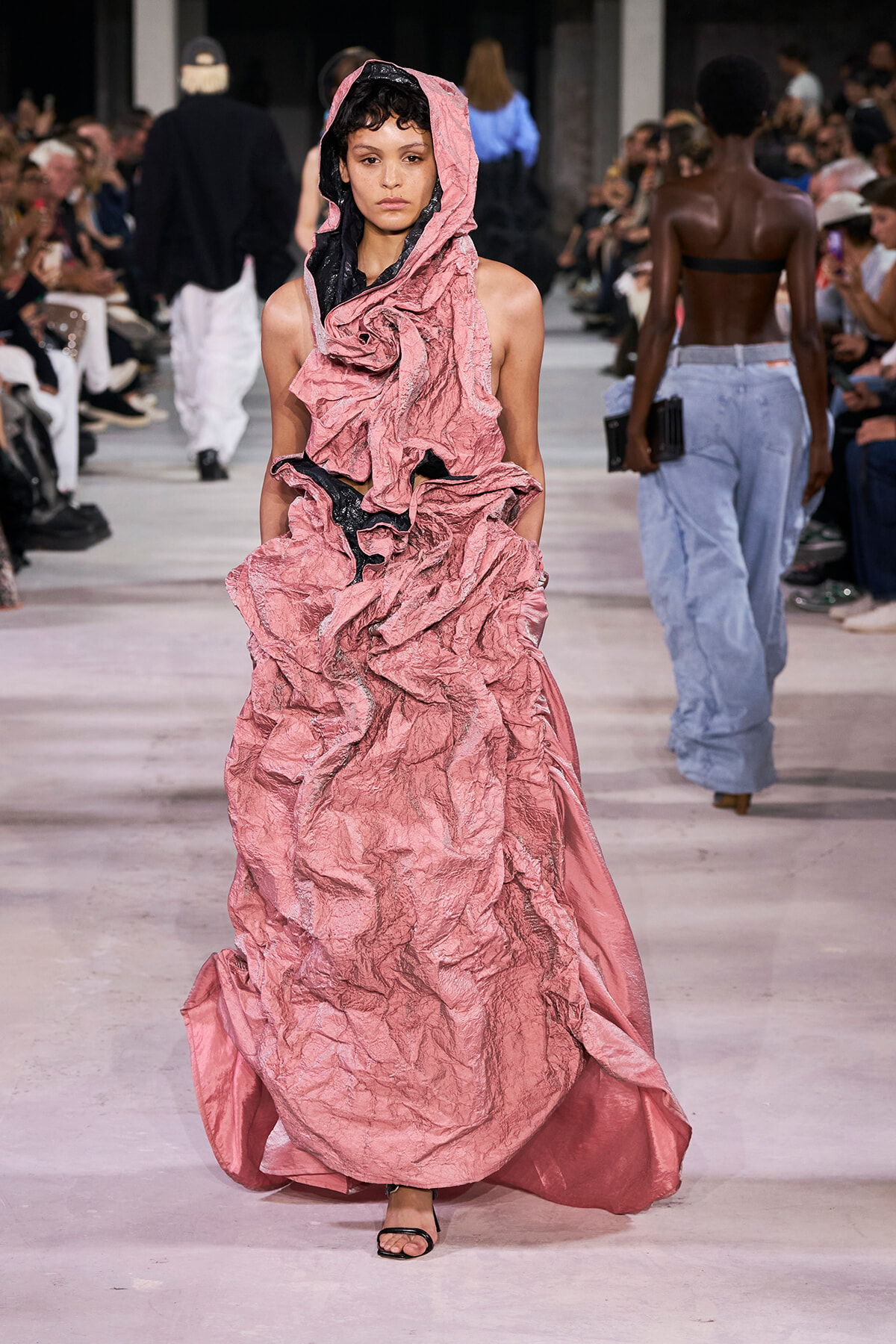 Model walking the runway in a voluminous pink crinkled gown with hood, black inner lining visible, audience on both sides.