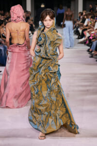 Model walks runway in a textured metallic gold-blue gown with a hooded scarf, while a pink dress-clad model faces away in the background; audience seated along the catwalk.