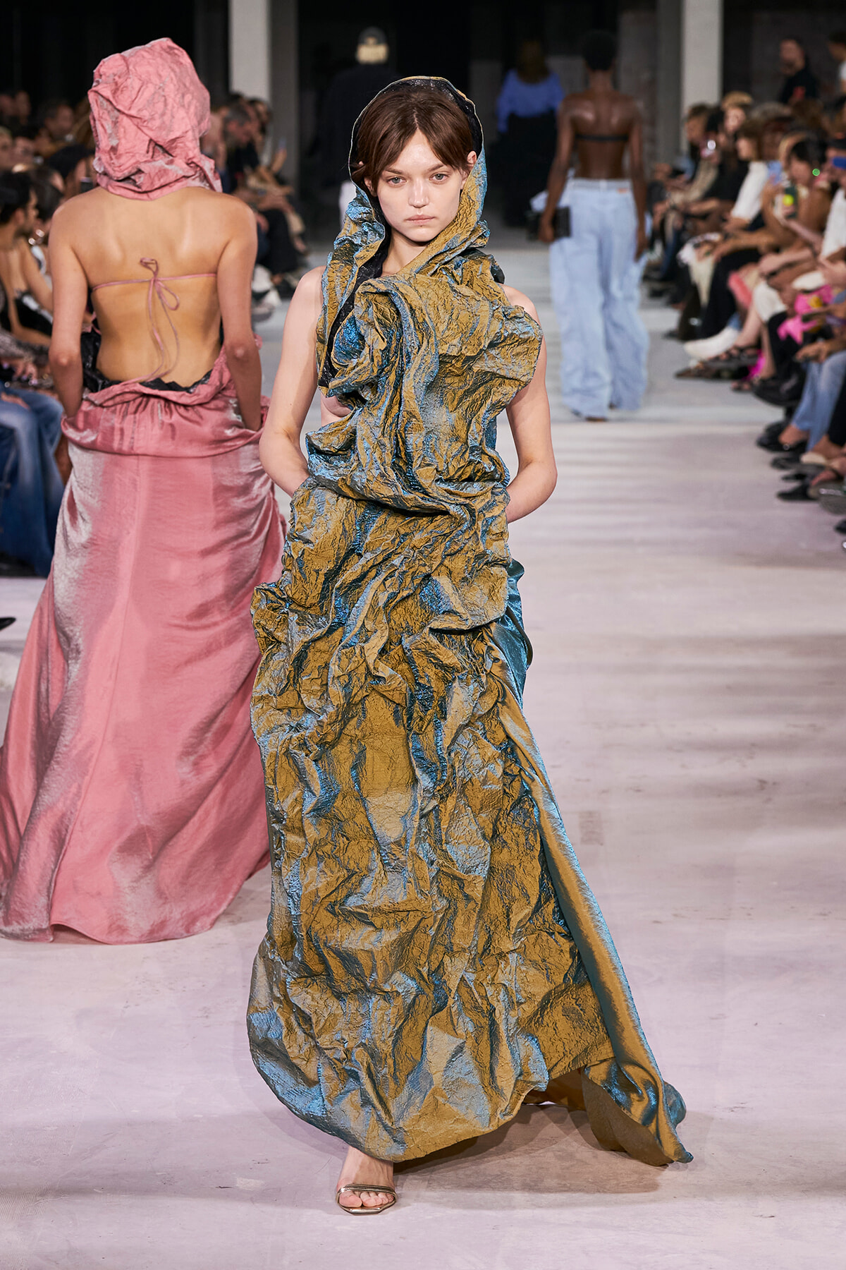 Model walks runway in a textured metallic gold-blue gown with a hooded scarf, while a pink dress-clad model faces away in the background; audience seated along the catwalk.
