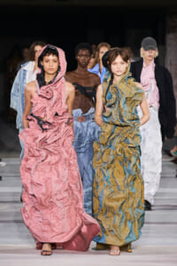 Models walk a runway wearing oversized crinkled gowns in pink, blue, and mustard colors.
