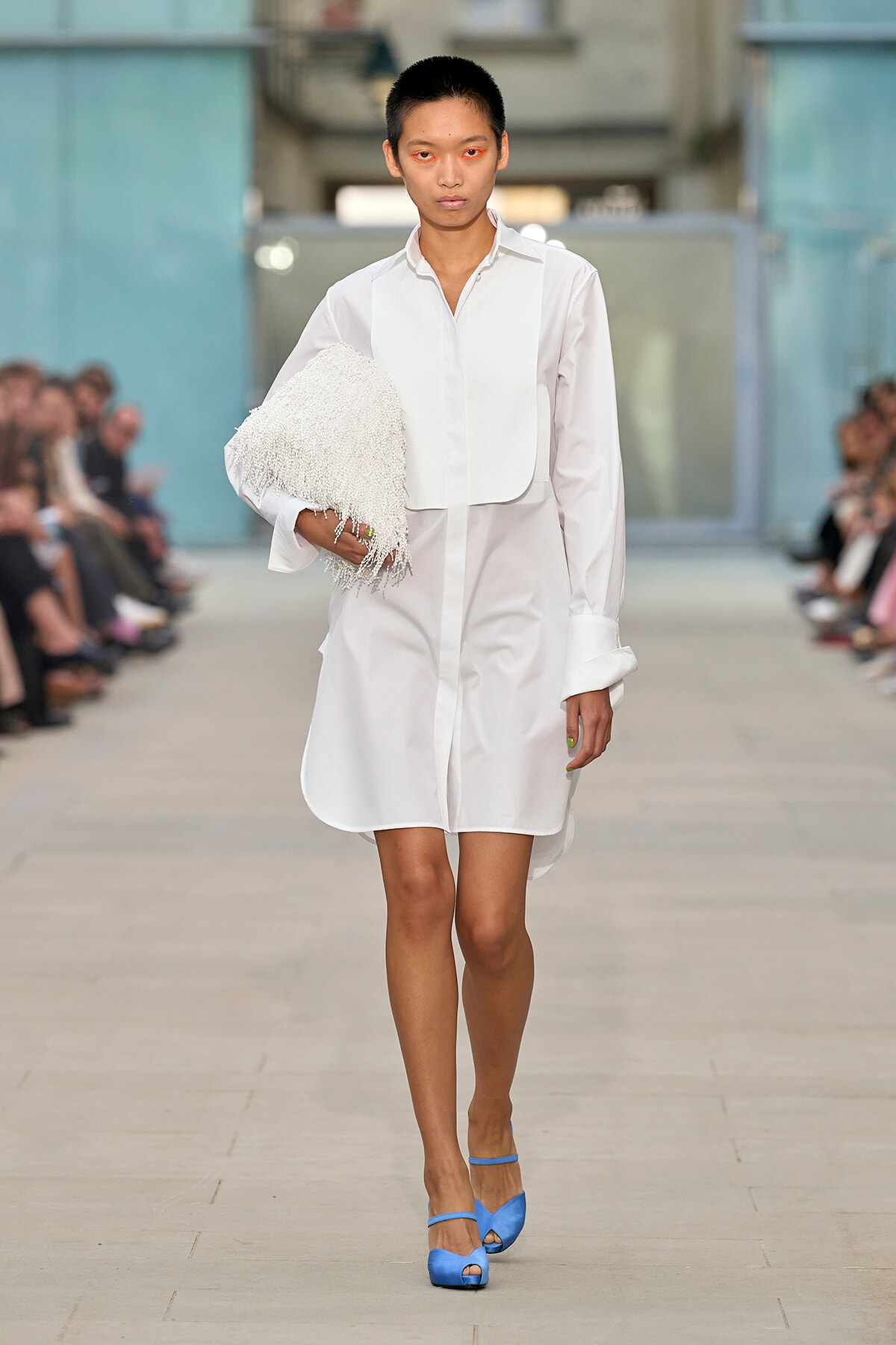 Model walking runway in a white shirt dress, holding a white fringed clutch and wearing blue heeled sandals.