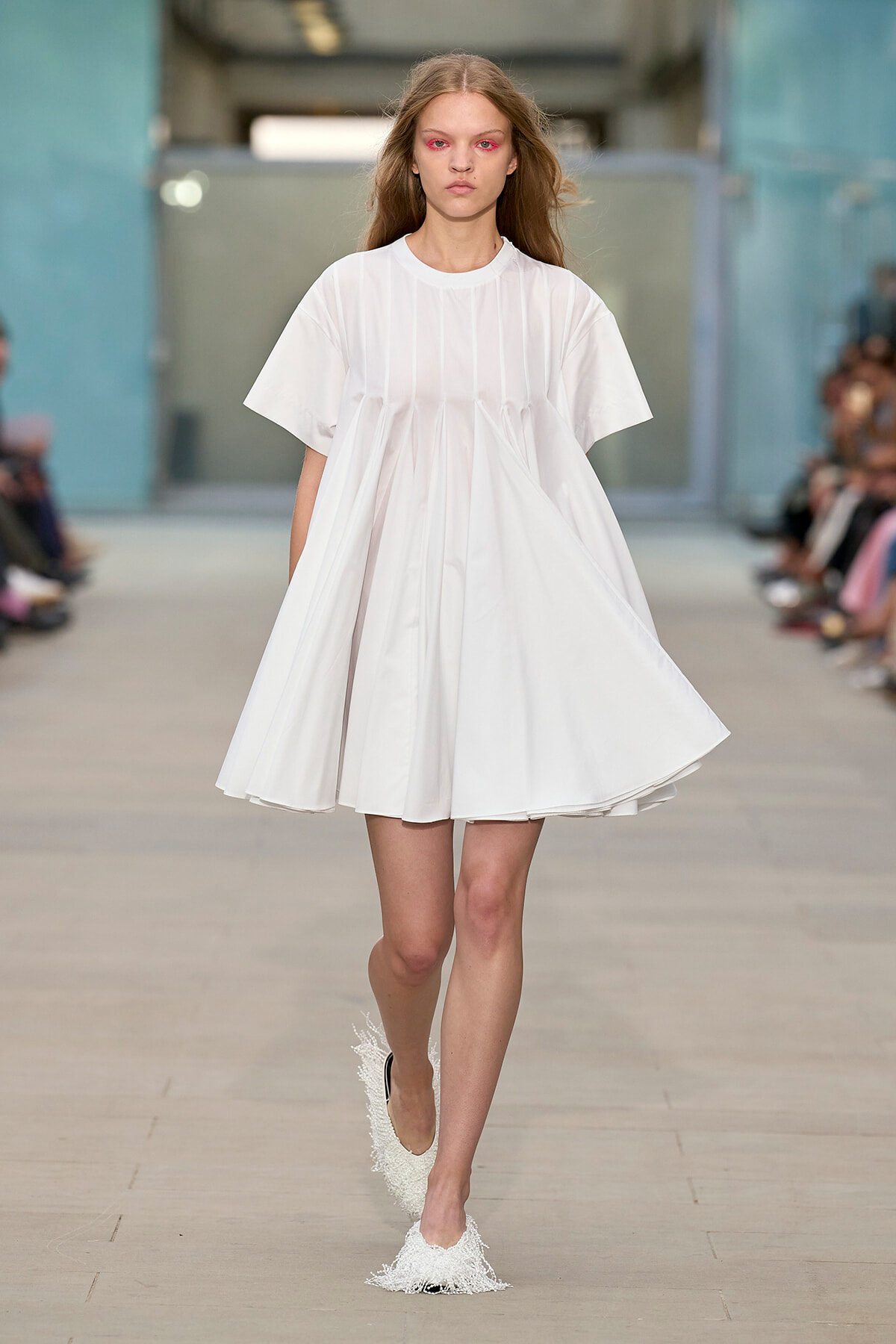 Runway model wearing a knee-length white dress with pleated panels walking toward the camera.