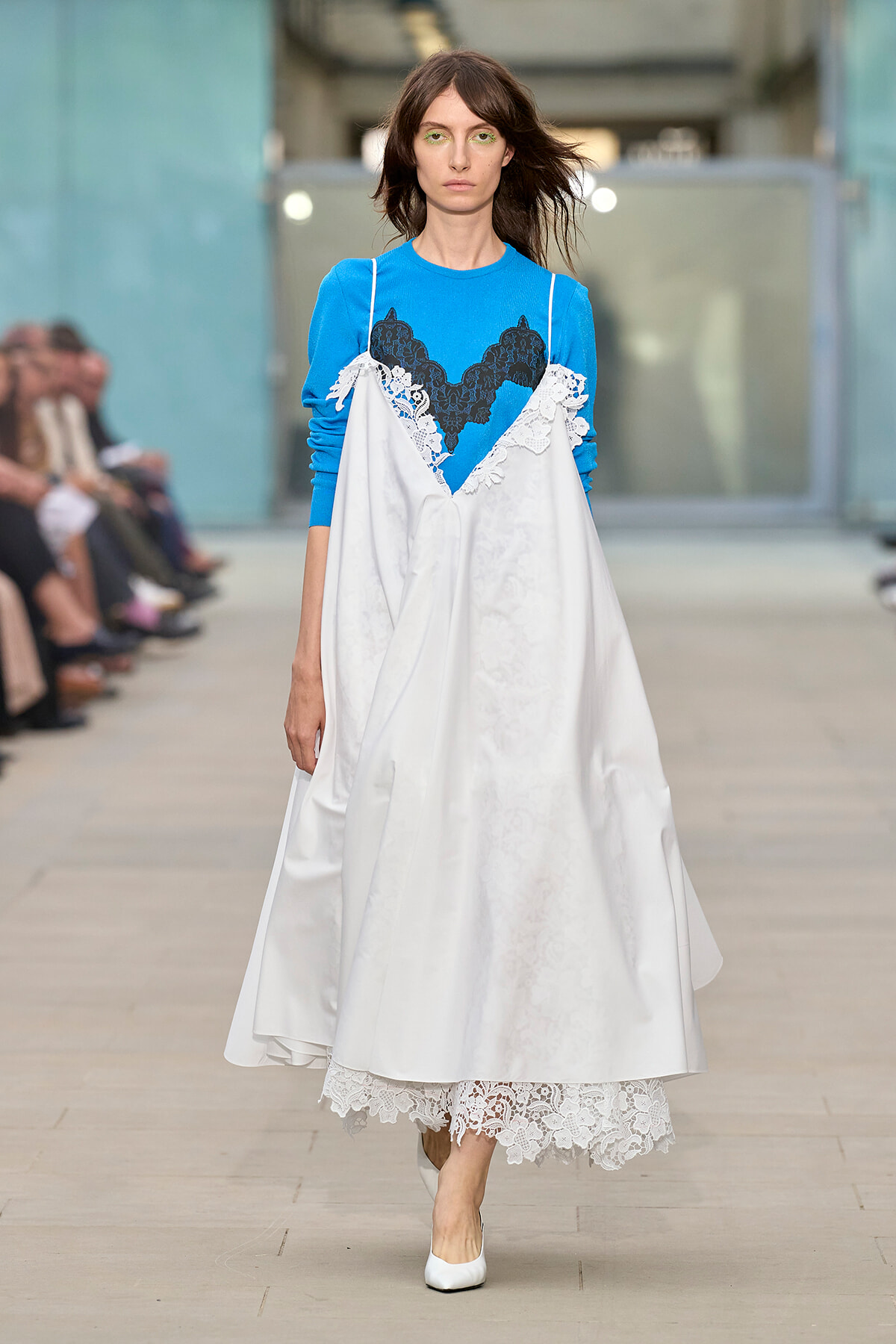 Model walks a fashion runway wearing a bright blue knit top under a white satin dress with lace trim and white heels.