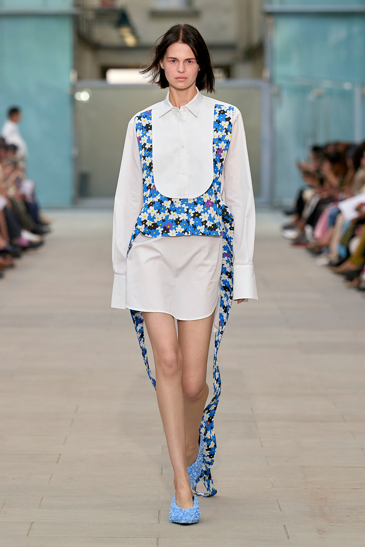 Model walking runway in white shirt layered with a blue floral vest and matching trailing fabric, wearing blue textured shoes.