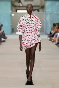 Runway model in a pink floral dress with voluminous sleeves walking down a catwalk, audience blurred in the background.