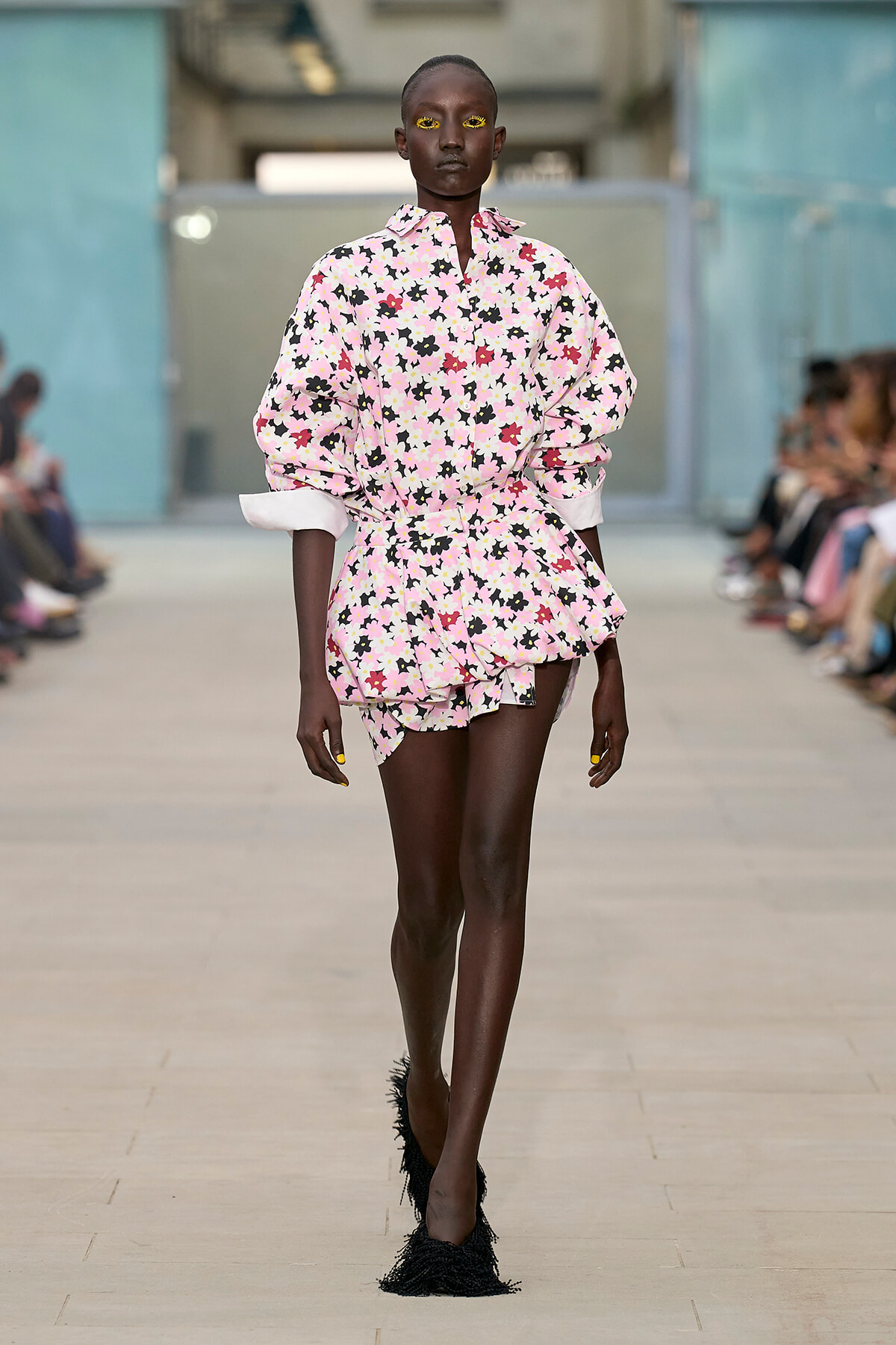 Runway model in a pink floral dress with voluminous sleeves walking down a catwalk, audience blurred in the background.