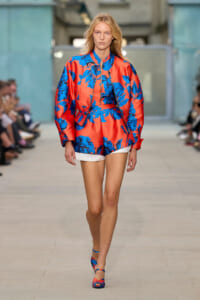 Model walking the runway in a bold red and blue floral satin bomber jacket with matching shorts; audience blurred in the background.