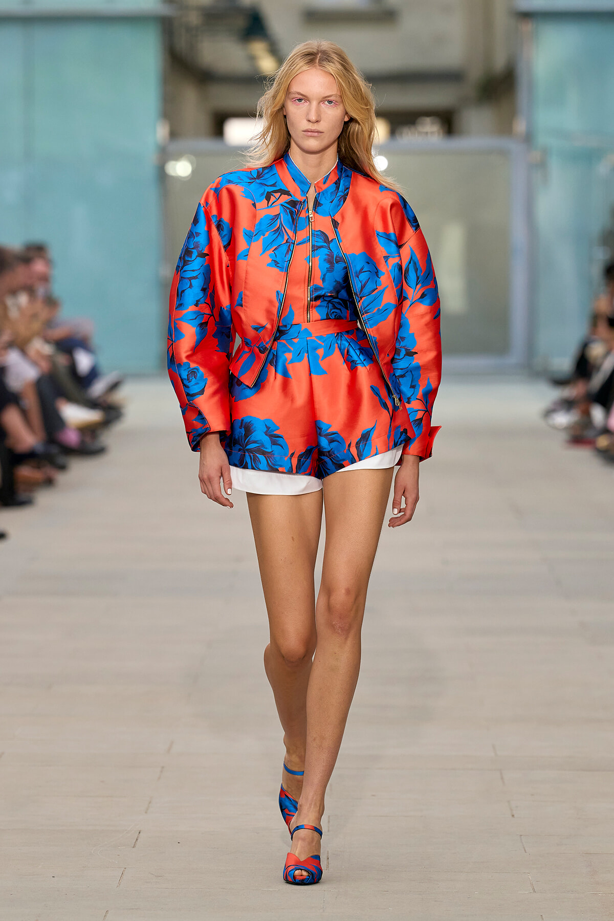 Model walking the runway in a bold red and blue floral satin bomber jacket with matching shorts; audience blurred in the background.