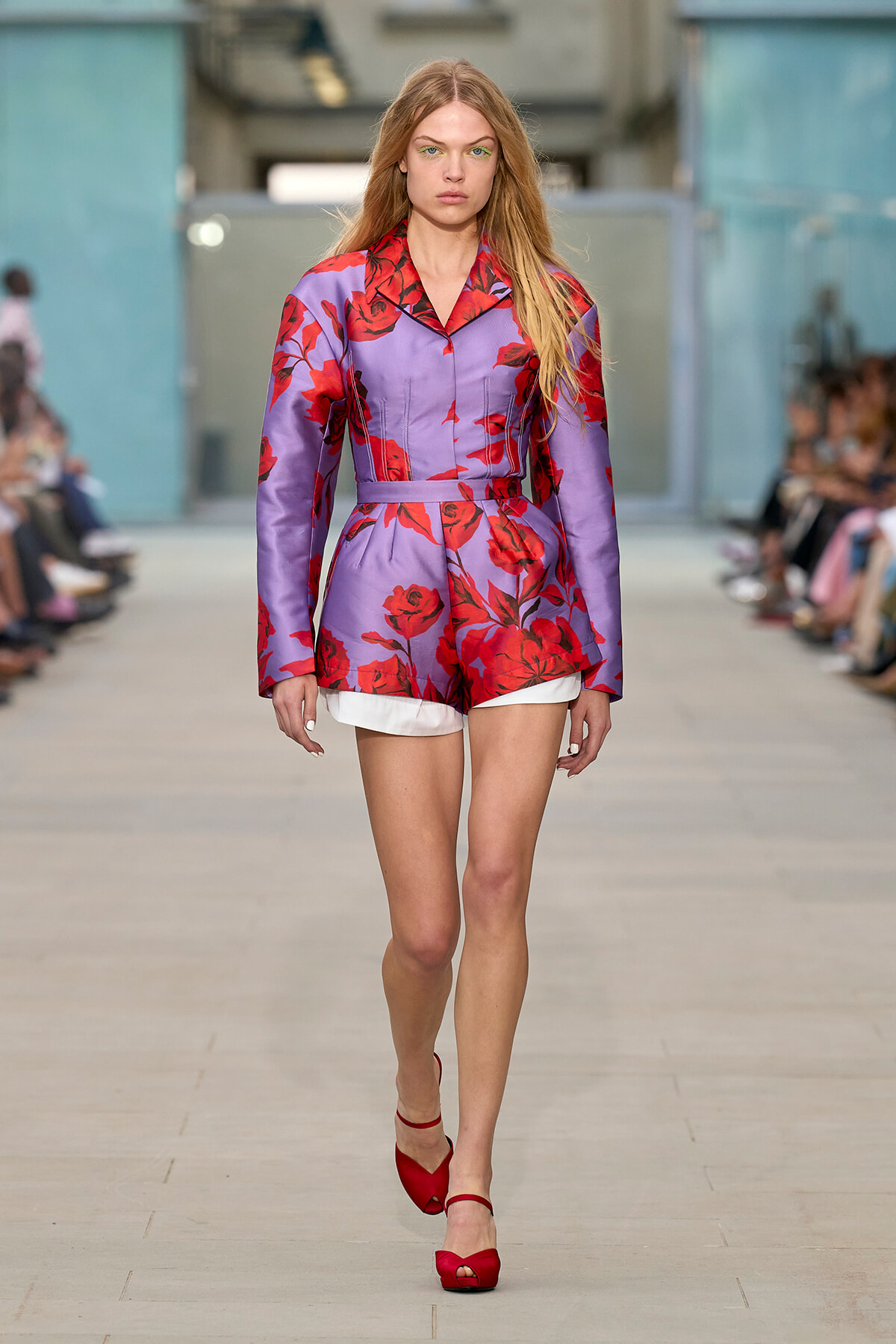 Model walking a runway in a lavender floral outfit with red roses, belted at the waist, white shorts peeking out, and red heeled sandals
