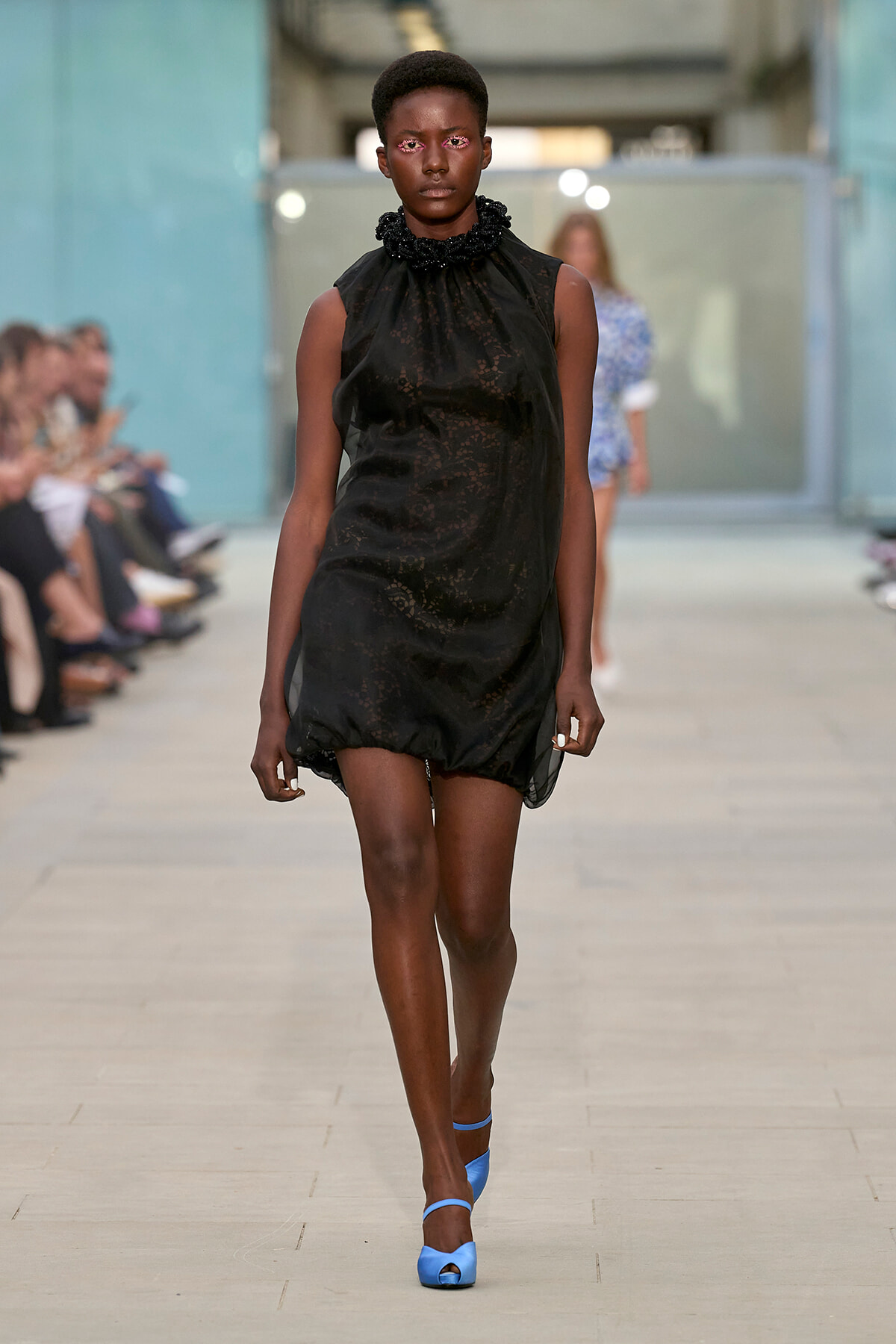 Model walking a runway in a black lace sleeveless dress with a ruffled high collar and bright blue strappy heels.