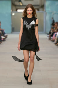 Model on a runway wearing a black sleeveless dress with white lace appliqué and sheer panel details, walking forward with a focused expression.