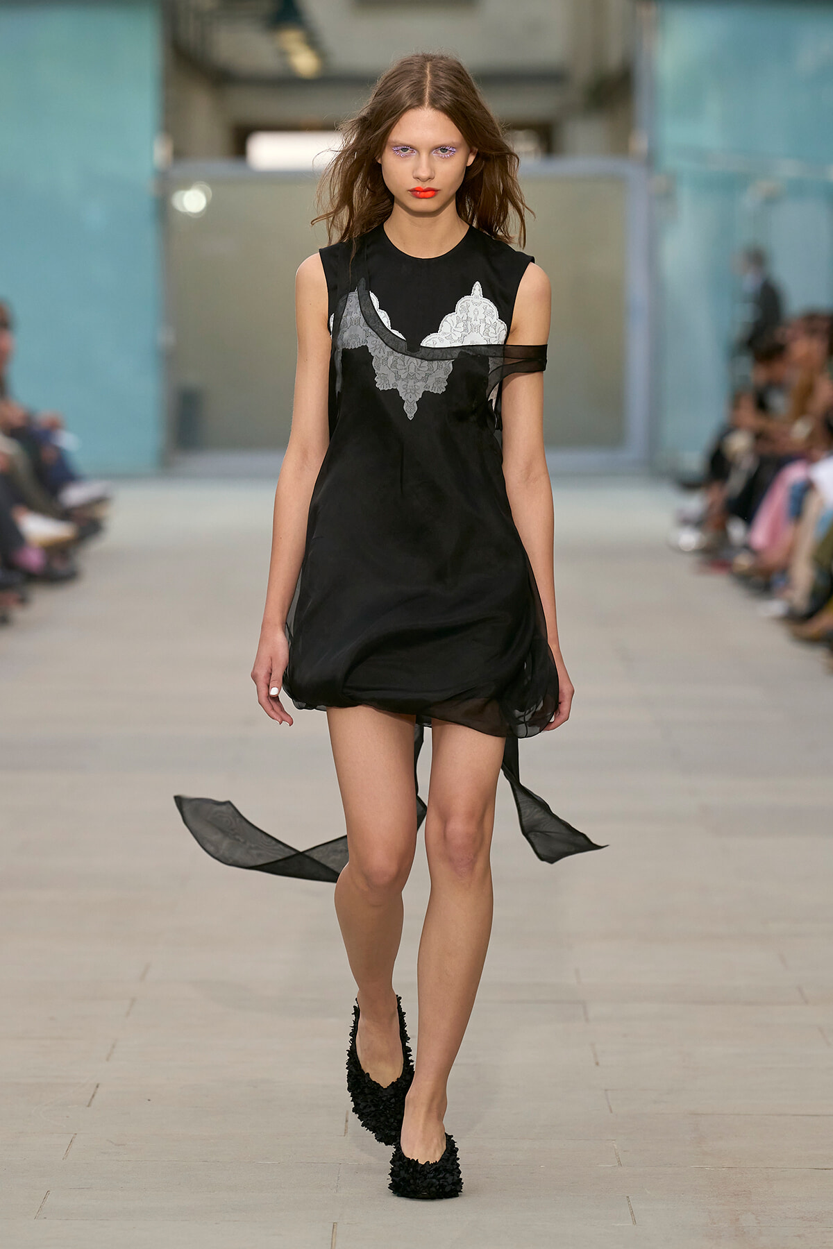 Model on a runway wearing a black sleeveless dress with white lace appliqué and sheer panel details, walking forward with a focused expression.