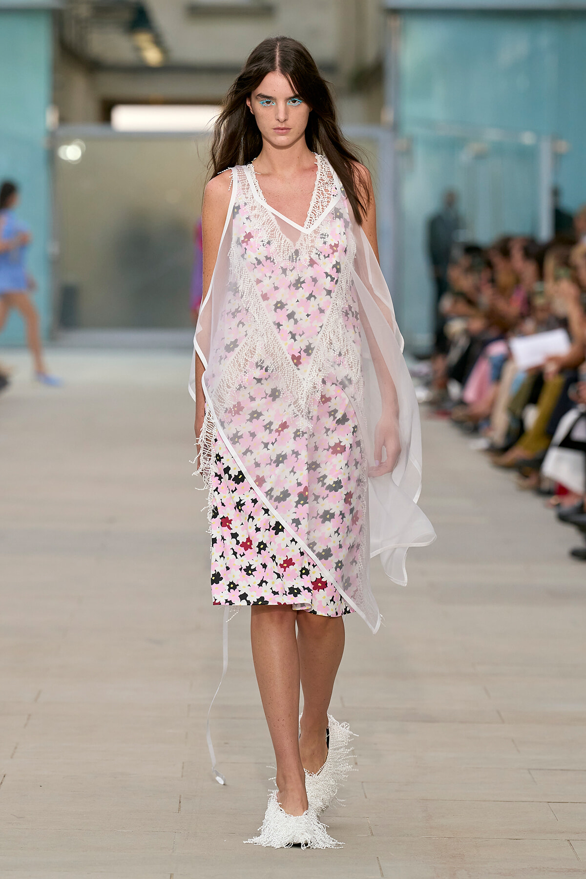 Model walking a runway in a pastel floral dress with a sheer white overlay and lace trim, audience blurred in the background, fashion show setting.