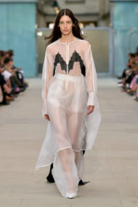 Model walks a runway in a sheer white dress with black lace panels, green eye makeup, and pale pink lipstick on a fashion show catwalk.