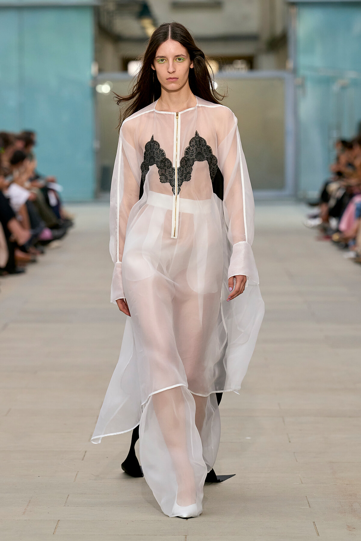 Model walks a runway in a sheer white dress with black lace panels, green eye makeup, and pale pink lipstick on a fashion show catwalk.