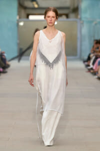 Model walks the runway in a sheer white sleeveless dress with a gray fringe chevron design at the bodice.