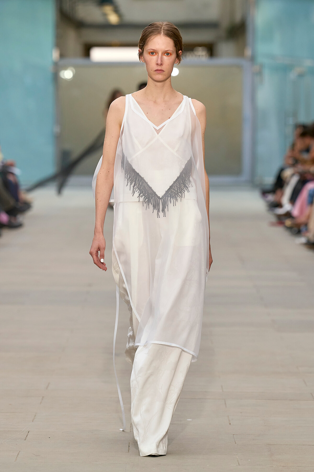Model walks the runway in a sheer white sleeveless dress with a gray fringe chevron design at the bodice.