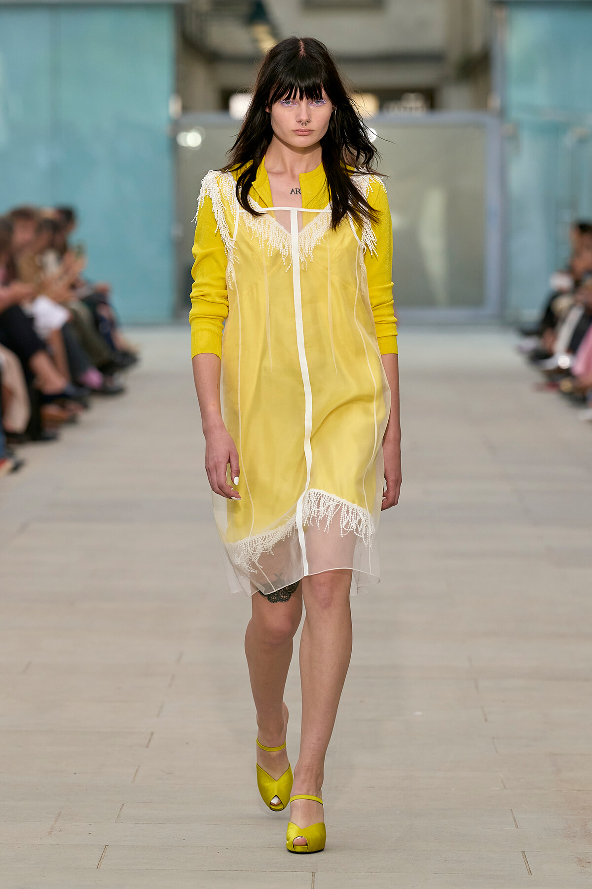 Model walking the runway in a yellow sheer dress with white fringe trim and matching yellow sandals