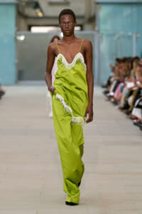 Model walks the runway in a lime-green slip jumpsuit with white beaded fringe, bright blue eye makeup, and an attentive pose as the audience watches.