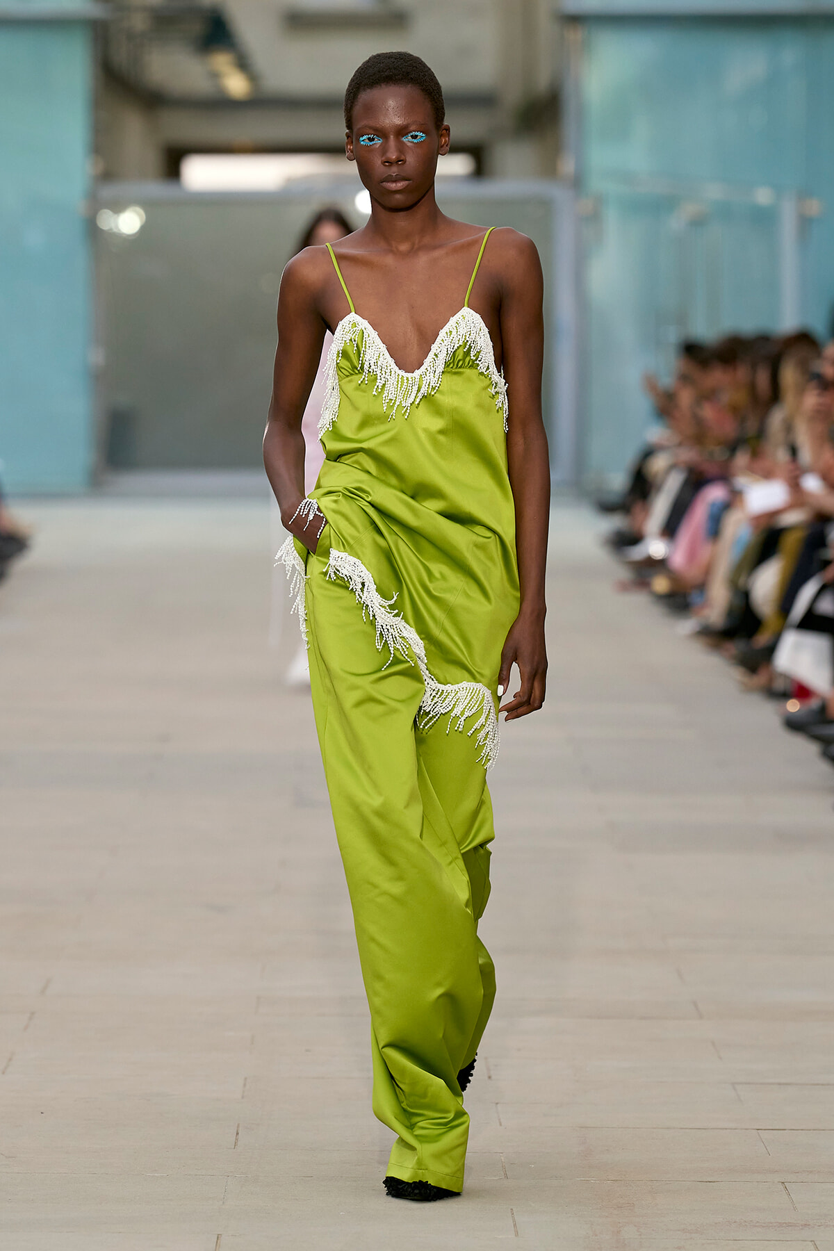 Model walks the runway in a lime-green slip jumpsuit with white beaded fringe, bright blue eye makeup, and an attentive pose as the audience watches.