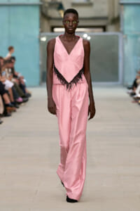 Model walks the runway in a pink satin sleeveless jumpsuit with a black fringe trim at the waist/torso area.