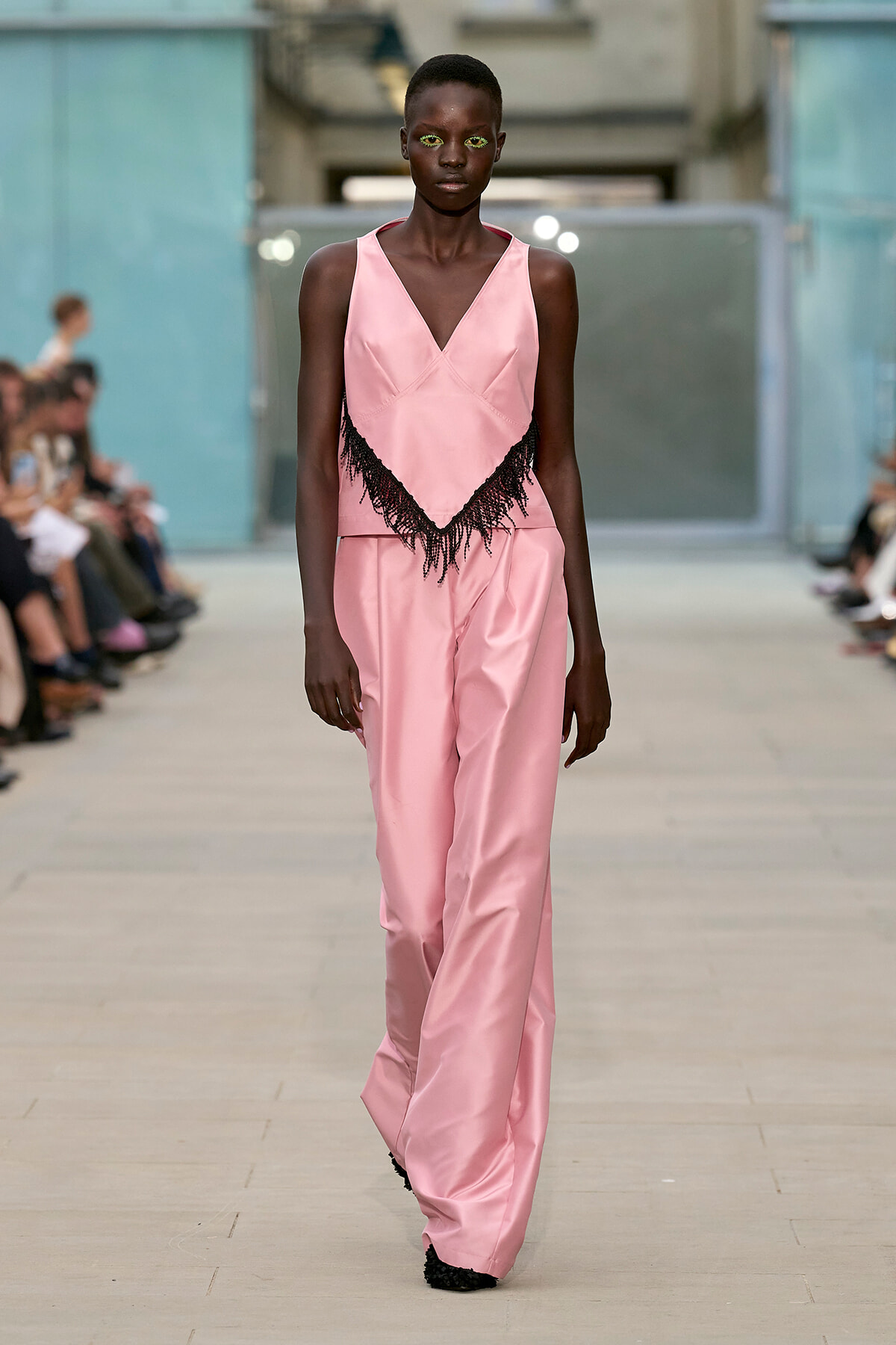 Model walks the runway in a pink satin sleeveless jumpsuit with a black fringe trim at the waist/torso area.