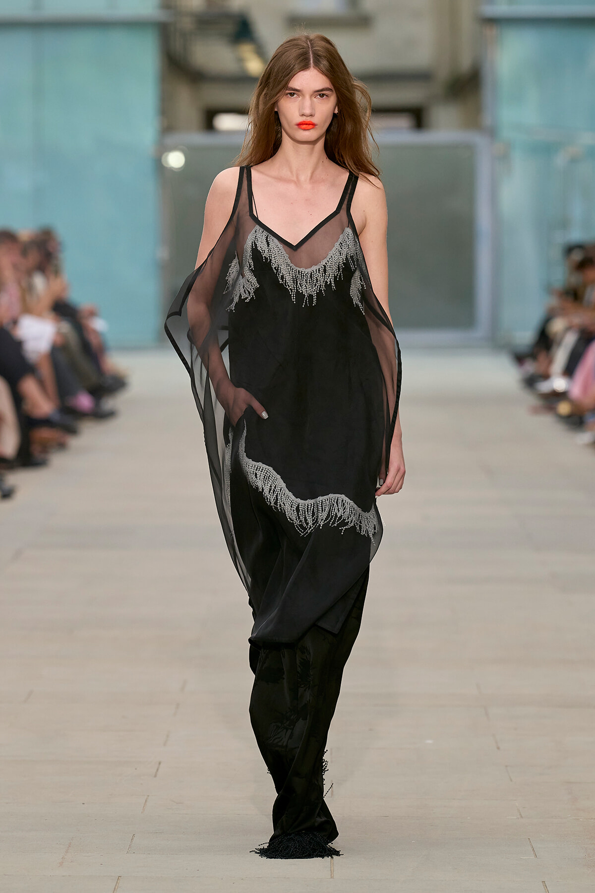 Model walking the runway in a black sheer dress with silver lace detailing at the neckline and hem.