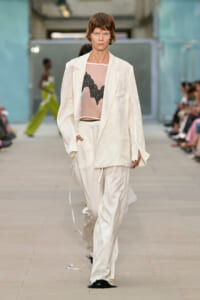 Model walks a runway wearing an oversized white suit with a sheer pink blouse and black lace bralette visible underneath. Audience blurred in the background.