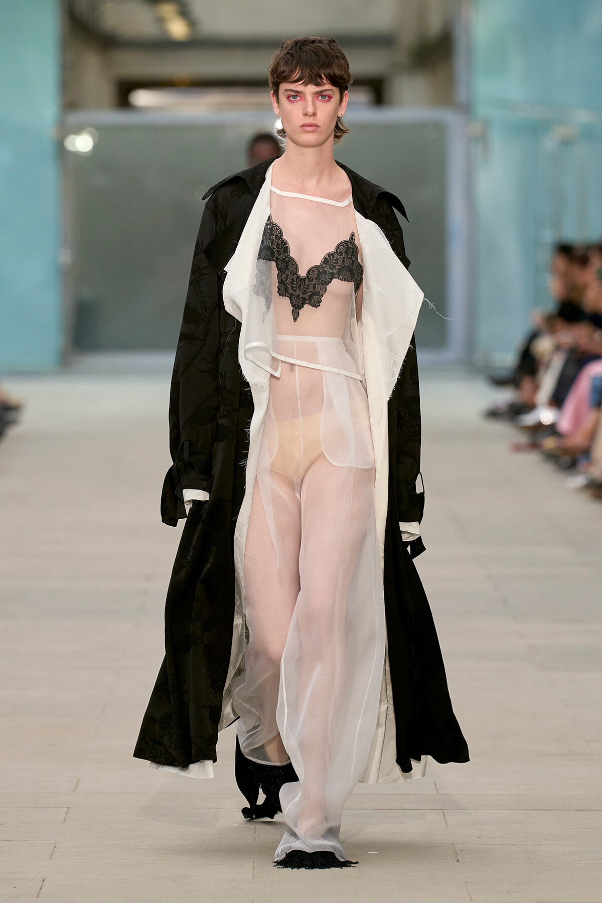 Model walks the runway in a sheer, pale pink dress with a black lace bralette, layered under a long black coat.