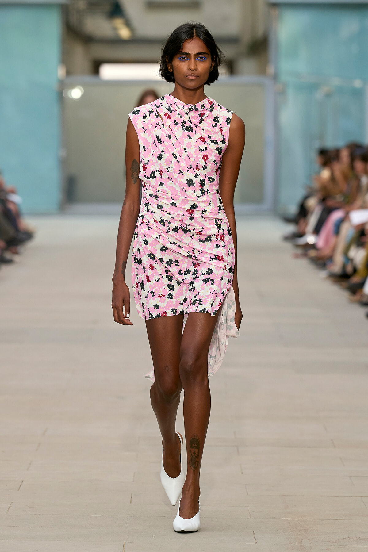 Model walks down fashion runway in a pink floral wrap dress with white heels, audience blurred in background.