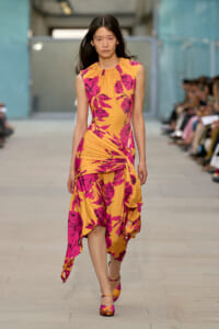 Model on a runway wearing a sleeveless orange dress with pink floral print and ruched detailing at the waist.