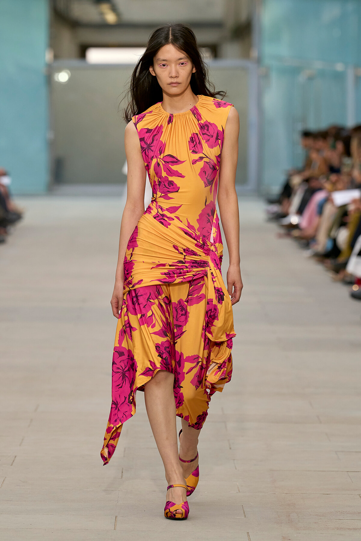 Model on a runway wearing a sleeveless orange dress with pink floral print and ruched detailing at the waist.