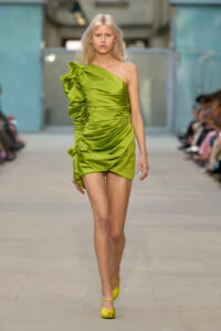Model walking the runway in a lime-green one-shoulder ruched dress with oversized ruffle detail, paired with yellow heels.