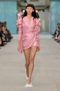 Model walks a fashion runway wearing a pink satin wrap mini dress with dramatic puff sleeves and ruching details.