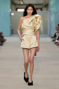 Model walking a runway in a cream satin one-shoulder top with a large bow and matching high-waisted shorts, black textured heels visible.