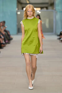 Model walking a fashion runway in a lime-green sleeveless dress with white lace trim at neck and hem, focused expression.