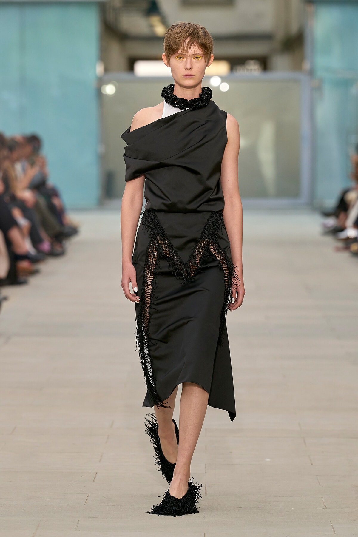 Model walking the runway in a black draped gown with fringe lace panels and a chunky black necklace.