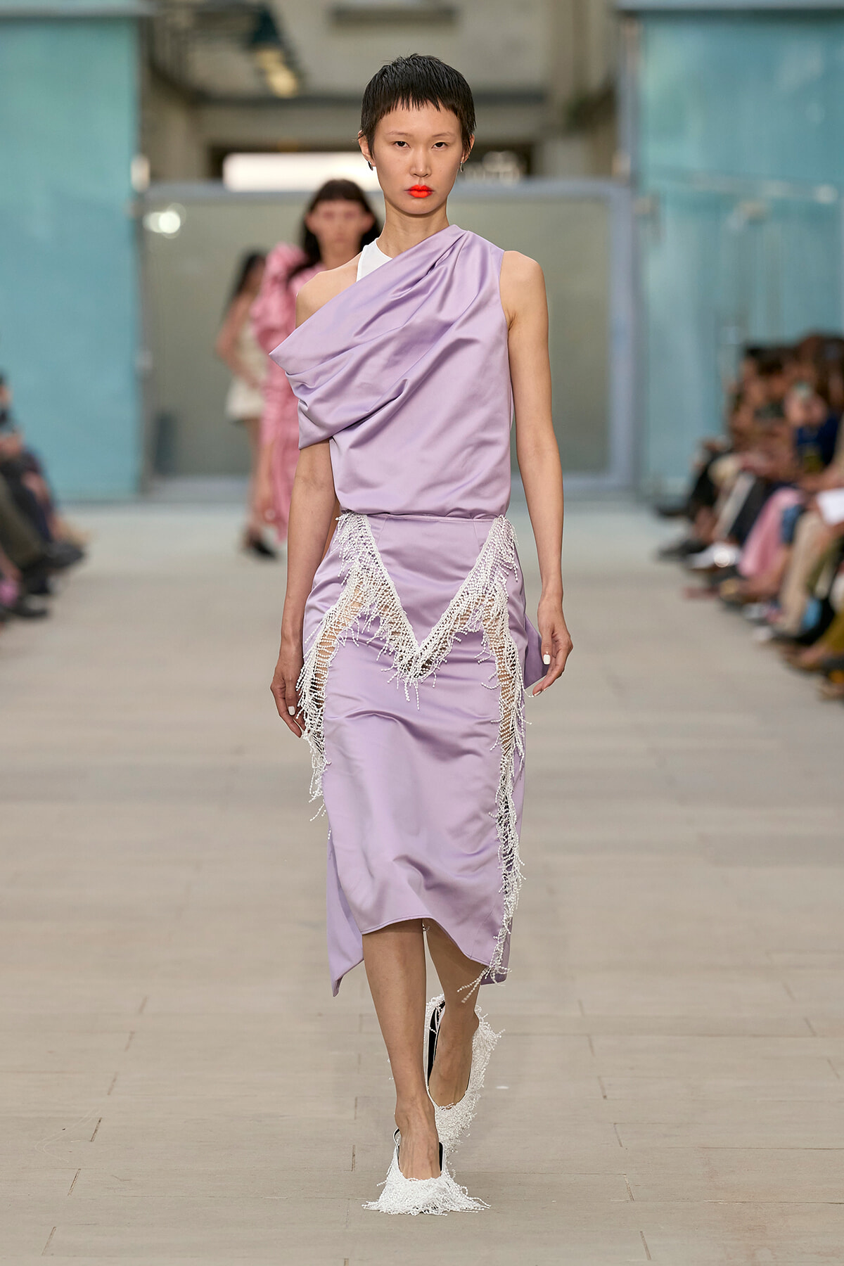 Model walks a fashion runway in a lavender satin dress with shoulder drape and white fringe detailing at the hem/waistline