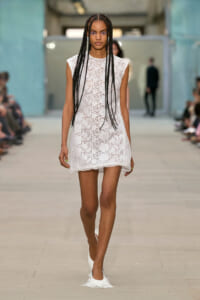 Model walks a fashion runway in a white lace sleeveless shift dress, with long box braids spilling over her shoulders.