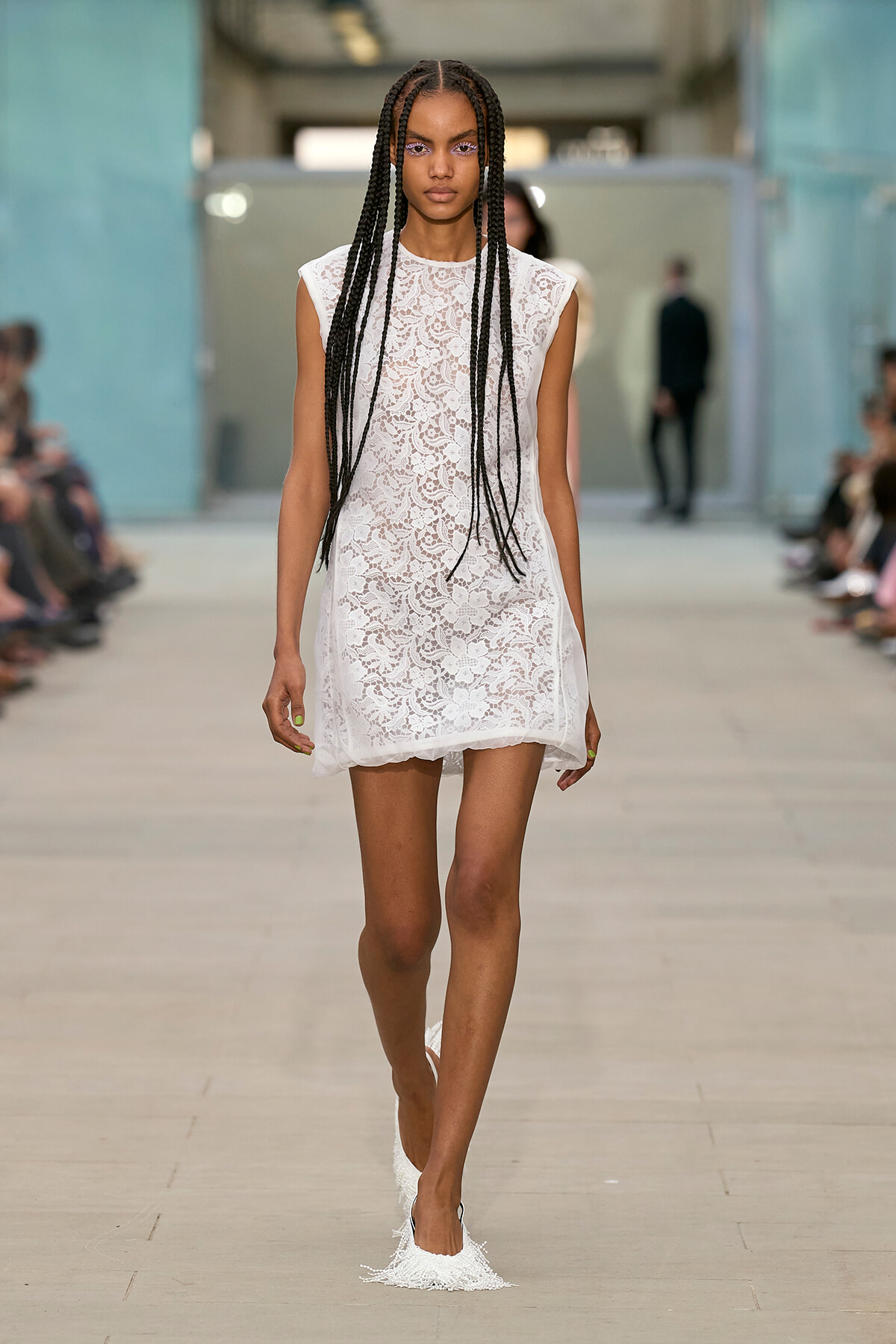 Model walks a fashion runway in a white lace sleeveless shift dress, with long box braids spilling over her shoulders.