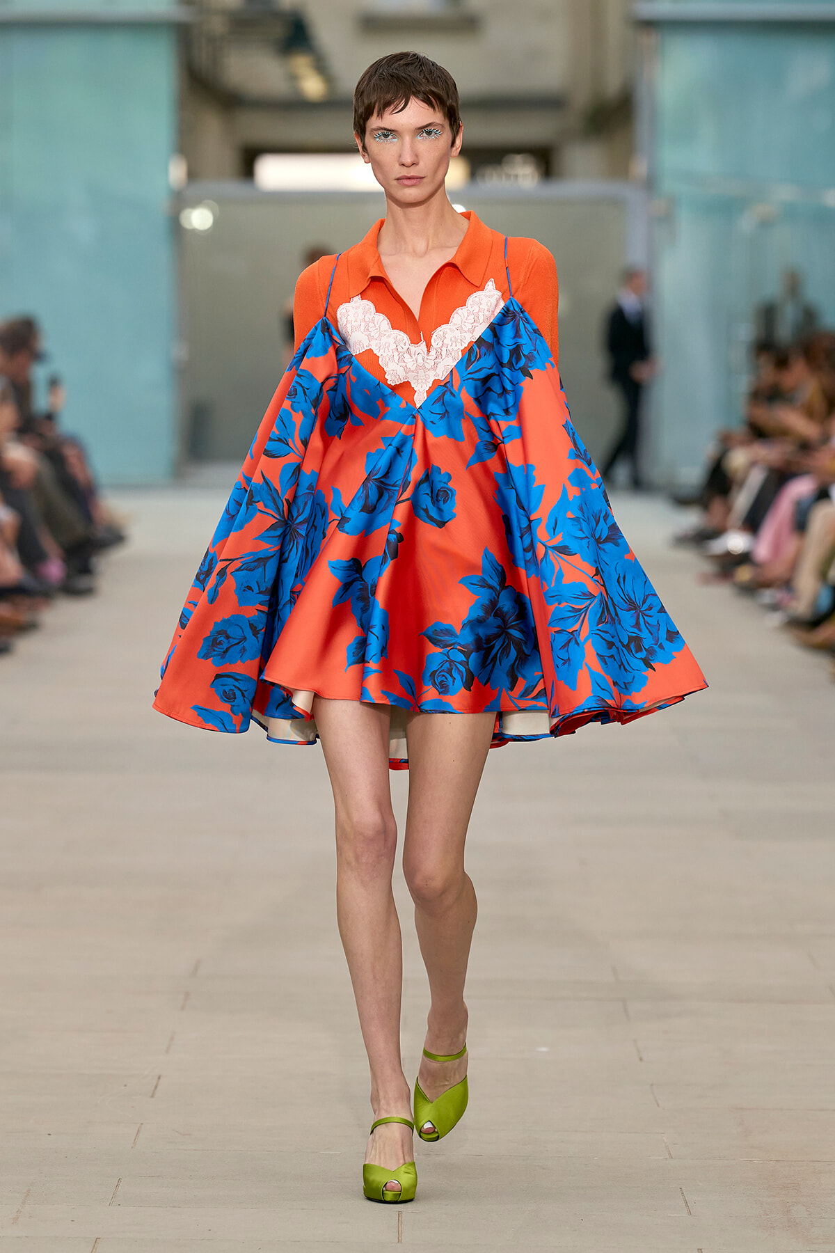 Model walking a runway in an orange and blue floral cape dress with white lace trim and lime-green heels, audience blurred in the background.