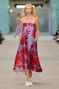 Model walking a runway in a strapless lavender gown with a bold red rose print and fluffy white shoes.