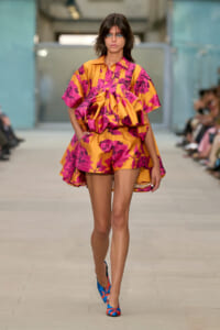 Model walking a runway in a vibrant orange and pink floral cape dress with matching shorts; blue floral-patterned heels visible, in a fashion show setting.