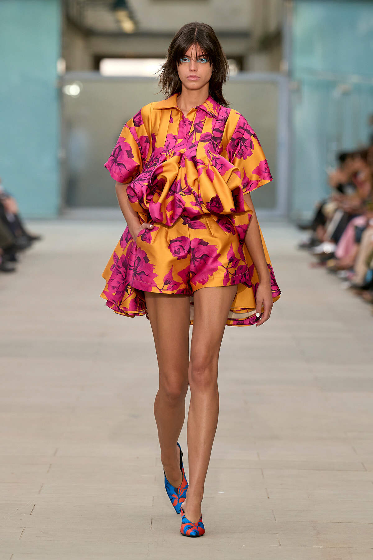 Model walking a runway in a vibrant orange and pink floral cape dress with matching shorts; blue floral-patterned heels visible, in a fashion show setting.