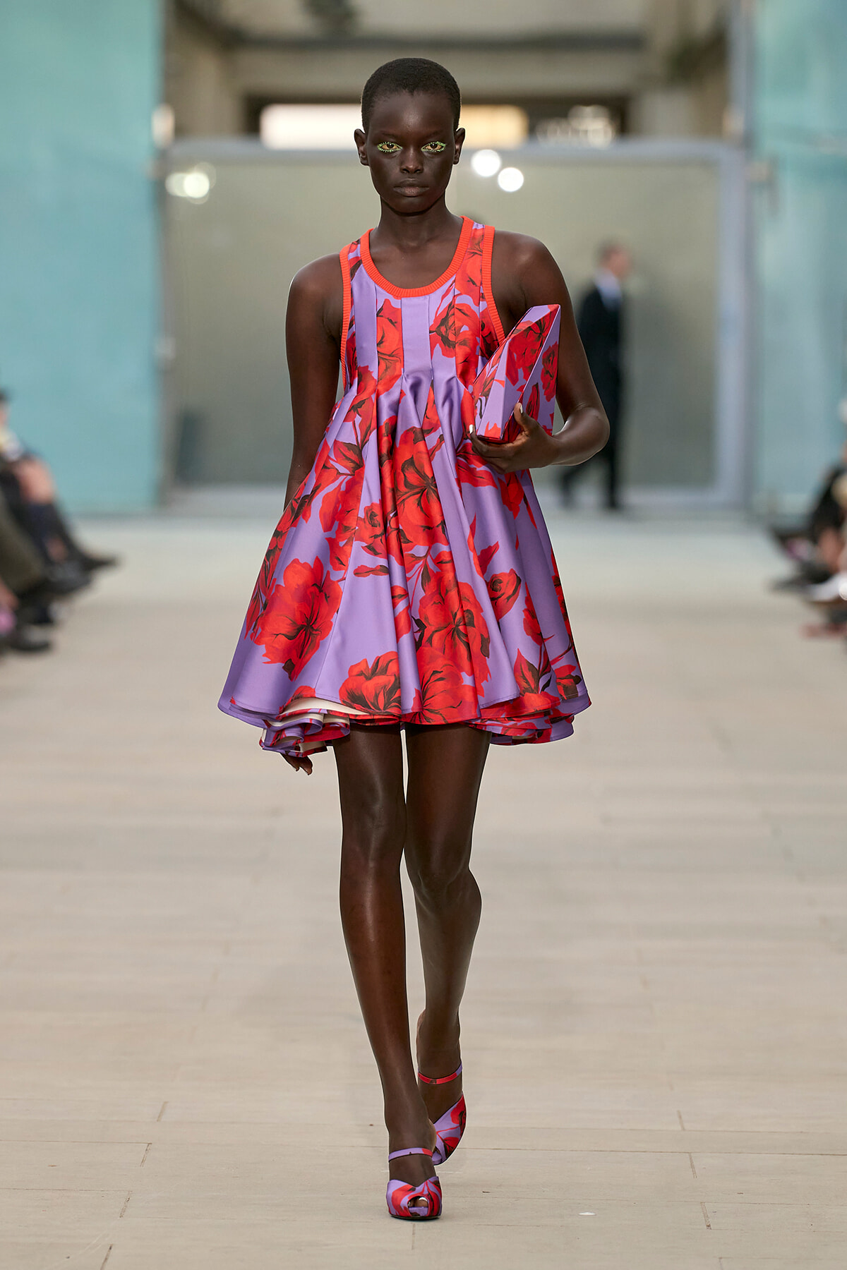 Model walks a runway in a lavender dress with red floral print, carrying a matching angular clutch.