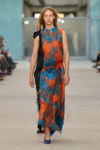 Model walking the runway in a sleeveless orange and blue fringe dress with a black side panel, paired with colorful heeled shoes.