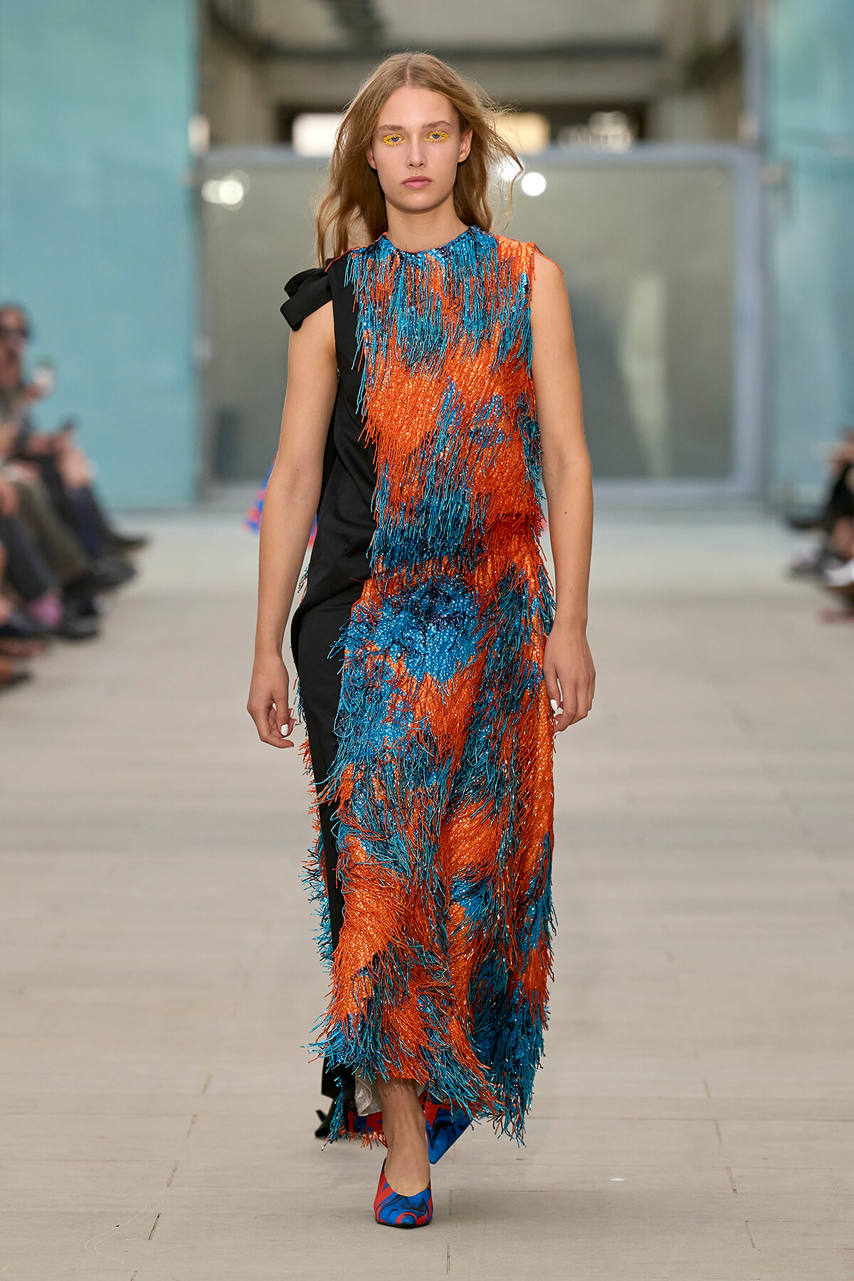 Model walking the runway in a sleeveless orange and blue fringe dress with a black side panel, paired with colorful heeled shoes.