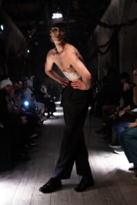 Male model on a dim runway, shirtless with a white belt and black wide-leg pants, hands on hips, cap, audience lining the sides.