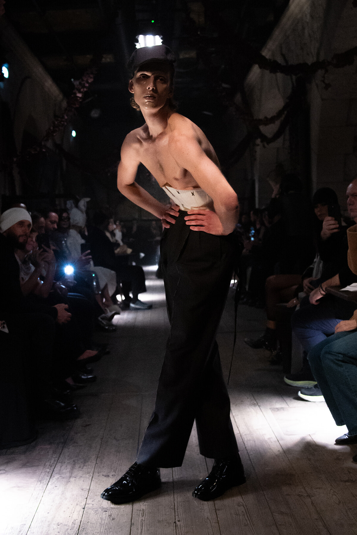 Male model on a dim runway, shirtless with a white belt and black wide-leg pants, hands on hips, cap, audience lining the sides.