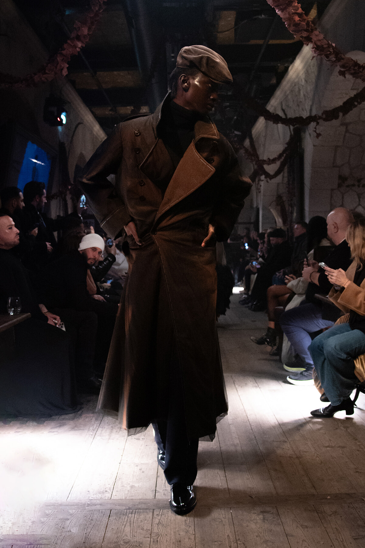Model on a fashion runways wearing a long brown leather coat and a newsboy cap, with audience seated along both sides of the wooden catwalk.